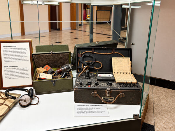 BC-221 frequency meter in its open wooden case beside its calibration book, headphones and a green accessory box, in a glass display