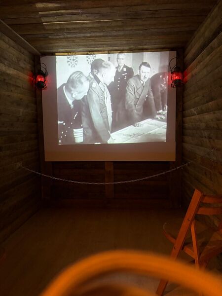 Small wood lined museum cinema showing black and white wartime film of officers studying a map