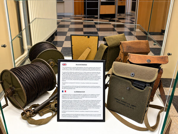 Field telephone equipment with large drums of telephone cable and canvas carrying bags, beside an information board in a glass case
