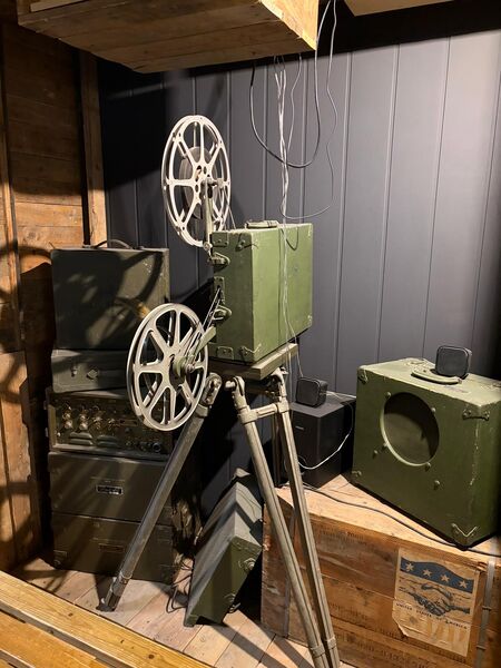 Wartime film projector with two reels on a tripod, beside green equipment cases and a speaker