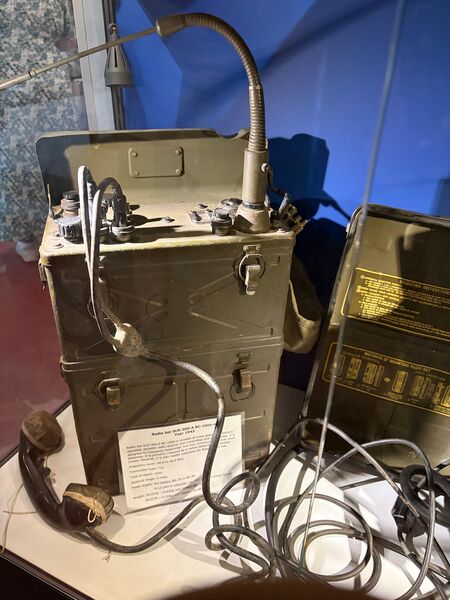 Olive green military radio set with a gooseneck microphone, headset and coiled cables, with an information card