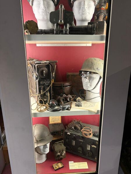 Tall glass display case with shelves of wartime radio sets, headsets and helmets on mannequin heads