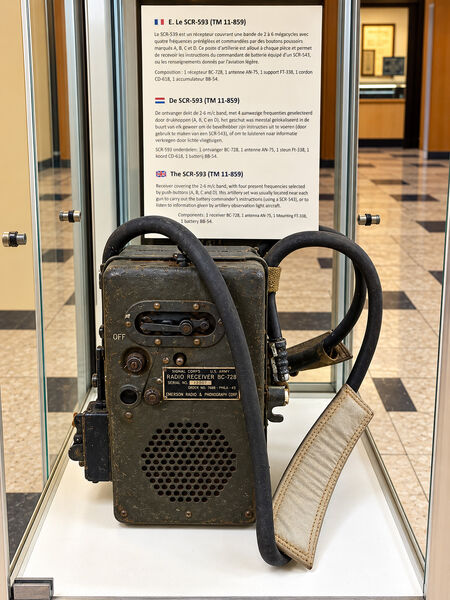 SCR-593 military radio receiver with a carrying strap and cable in a glass case, with an information board behind