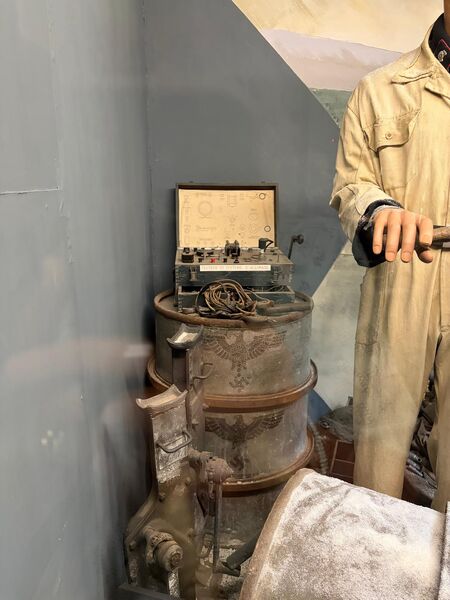 Small spy radio set in an open case, resting on top of a rusty drum next to a mannequin in a pale coat