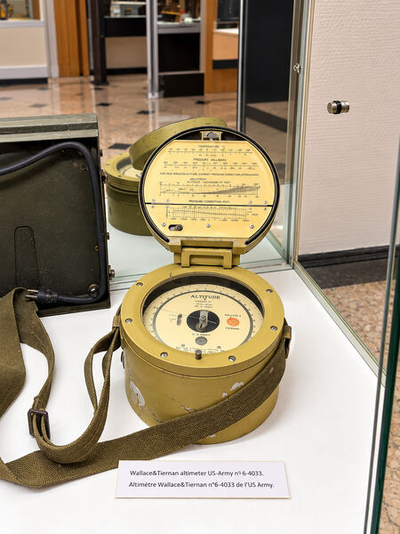Wallace and Tiernan US Army altimeter in an open round case with a carrying strap, in a glass display