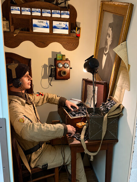 Museum diorama of a mannequin soldier wearing headphones at a desk of radio equipment, with a wall telephone and a framed portrait behind